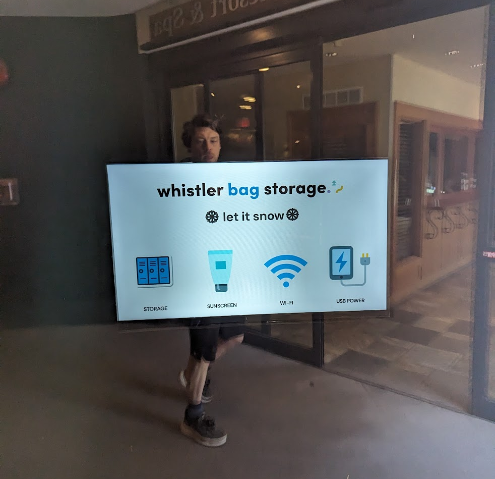 Person in reflection looking at tv that reads “whistler bag storage. let it snow” with icons for storage, sunscreen, Wi-Fi, and USB power.
