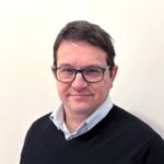 A man with short dark hair and glasses wearing a light blue collared shirt and dark sweater stands in front of a plain light-colored background.