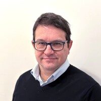 A man with short dark hair and glasses wearing a light blue collared shirt and dark sweater stands in front of a plain light-colored background.