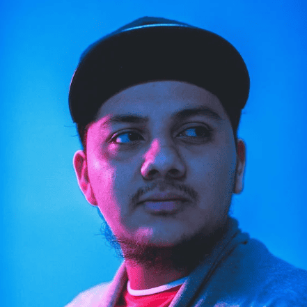 A person wearing a hat and jacket looks off to the side under blue and pink lighting.